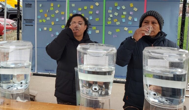 Two people in black jackets drink from small glasses as they stand behind large, clear containers of water. They are sampling recycled wastewater as part of an interactive exhibit.
