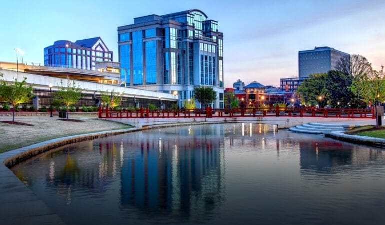 The serene Huntsville, Alabama, cityscape at dusk featuring a reflective canal bordered by a curved stone walkway and a red pedestrian bridge. Modern office buildings with glass facades and traditional brick buildings line the background, while lampposts and young trees add charm to the landscaped waterfront area. The soft evening sky casts a gentle glow over the scene.